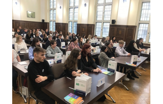 W przestronnej, jasnej sali o wysokich oknach i drewnianych panelach zgromadziła się duża grupa młodych ludzi. Siedzą w rzędach przy stołach, na których ustawiono torby z logo instytucji oraz materiały edukacyjne. Wśród uczestników panuje skupienie, a ich elegancki strój sugeruje oficjalny charakter wydarzenia. Numery na stolikach wskazują na organizację konkursu lub szkolenia.