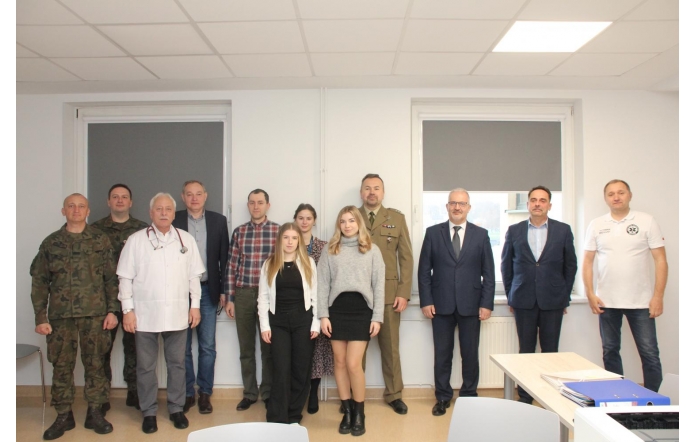 Grupa osób stoi wspólnie w jasnym pomieszczeniu na tle dwóch dużych okien z roletami. Wśród nich są żołnierze w mundurach, lekarz w białym kitlu, mężczyźni w garniturach i kobiety ubrane elegancko. Wszyscy patrzą w obiektyw.