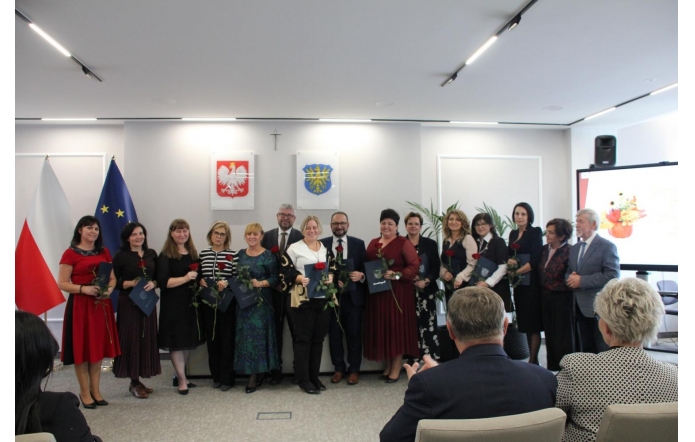 Grupa osób pozuje w sali sesyjnej. Uczestnicy trzymają róże i teczki, a w tle widoczne są flagi, godło Polski oraz herb Powiatu Cieszyńskiego.