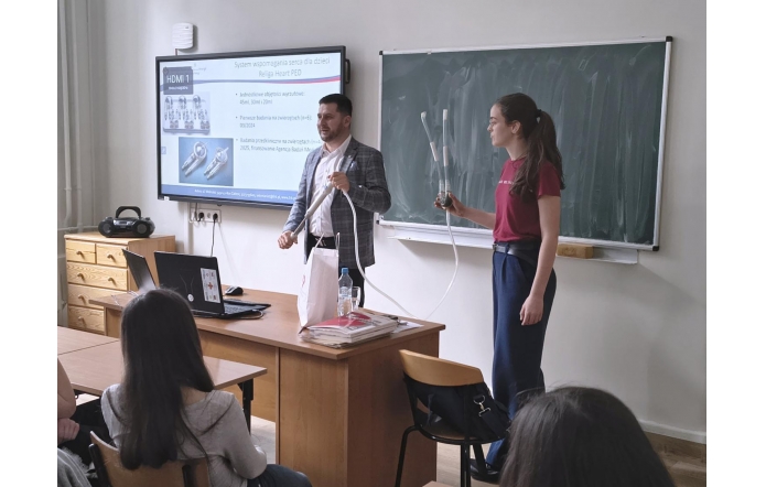 Sala wykładowa z tablicą kredową i ekranem prezentacyjnym. Mężczyzna i młoda dziewczyna stoją przy biurku; oboje trzymają elementy z przezroczystymi rurkami. Na ekranie wyświetlona jest prezentacja z ilustracjami urządzeń medycznych i tekstem. Przed nimi siedzą osoby skierowane w stronę prowadzących. Na biurku znajduje się laptop, butelka wody i stos dokumentów.