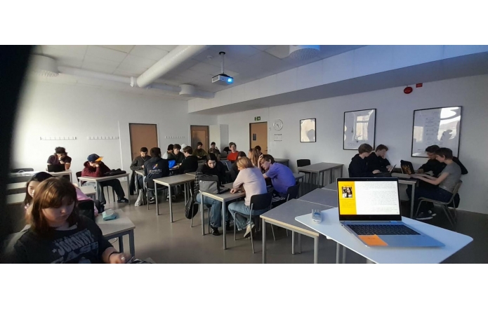 Uczniowie pracujący w grupach przy stolikach, na biurkach laptopy, z przodu otwarty komputer i projektor pod sufitem.