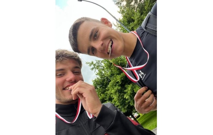 Dwóch młodych mężczyzn w sportowych strojach uśmiecha się szeroko, trzymając w ustach medale zawieszone na biało-czerwonych wstążkach. W tle widać drzewa i fragment latarni ulicznej.