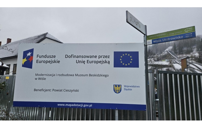 Tablica informacyjna o dofinansowaniu z Unii Europejskiej ustawiona przy ogrodzeniu, obok znak kierunkowy i zabudowania na tle ośnieżonego zbocza.