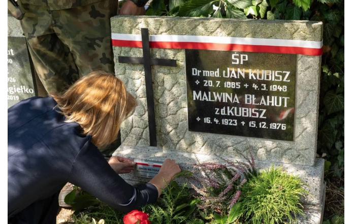 Kobieta przy grobie ozdobionym biało-czerwoną wstęgą umieszcza oznaczenie na płycie nagrobnej. Wokół widoczne są rośliny oraz kwiaty.