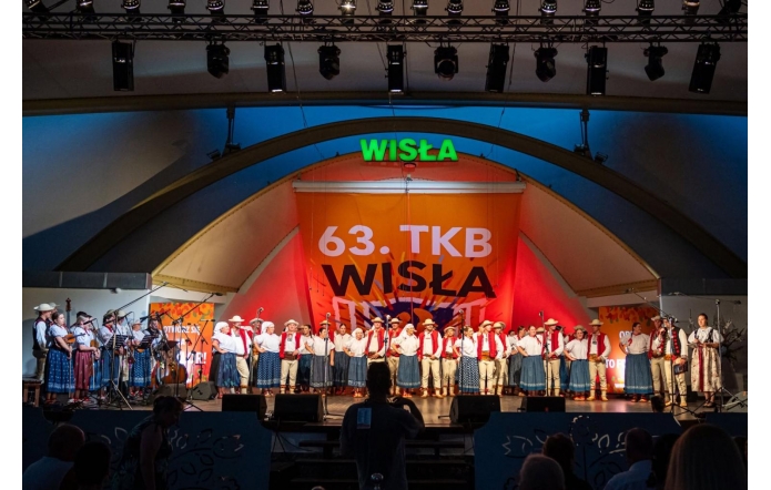 Grupa osób w strojach regionalnych stoi w rzędzie na scenie. Po lewej stronie widoczni są muzycy z instrumentami, a po prawej dwie kobiety stoją przy mikrofonach. W tle znajduje się napis „63. TKB Wisła”. Oświetlenie sali jest ciemne, a światło sceniczne pada głównie na stojące osoby.