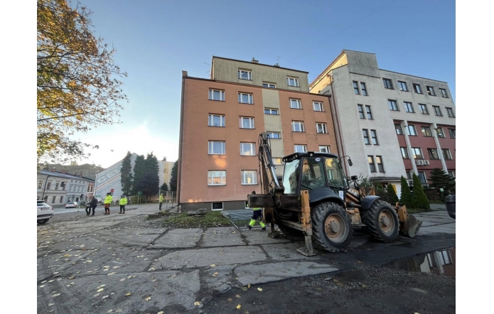 Na placu przed budynkiem trwa remont. Widoczna jest koparko-ładowarka, pracownicy w odzieży odblaskowej oraz fragment zabezpieczenia terenu robót. W tle znajduje się kilkupiętrowy budynek i sąsiednia zabudowa.