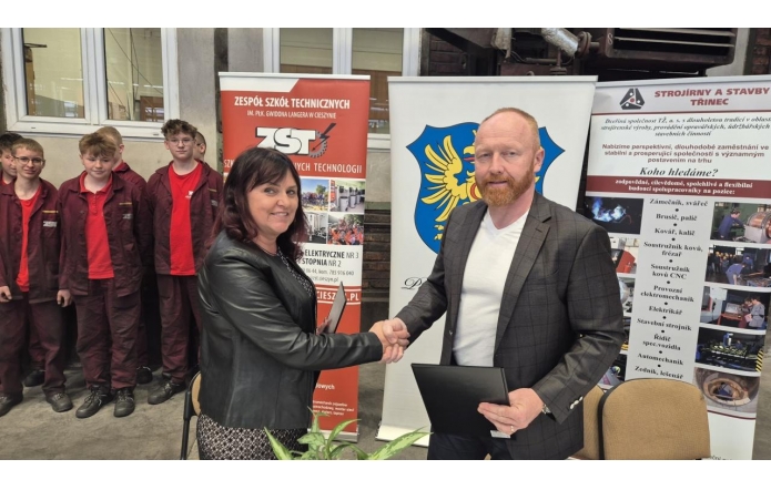 Bożena Cholewa, dyrektor Zespołu Szkół Technicznych im. płk. Gwido Langera w Cieszynie i Marian Czyż, prezes spółki Stroirny a stavby, uśmiechnięci uściskują dłonie. W tle grupa uczniów w czerwonych uniformach i roll-upy z logami obu instytucji.
