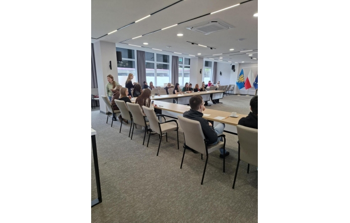 Sala sesyjna z ustawionymi w kształt litery U stołami i krzesłami. Przy stołach siedzi kilkunastu uczniów, część stoi. W tle widoczne są okna oraz ściana z flagami i logo na jasnym tle.