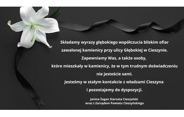 Czarne tło z delikatnym czarnym wstążkowym akcentem i białym kwiatem lilii w lewym górnym rogu. Na środku znajduje się tekst wyrażający kondolencje dla bliskich ofiar zawalenia kamienicy w Cieszynie, podpisany przez starostę oraz Zarząd Powiatu Cieszyńskiego. Prosty, elegancki styl.