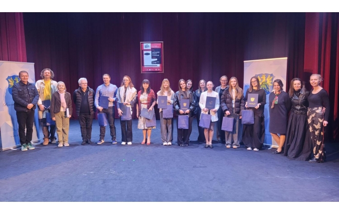 Grupa laureatów i organizatorów stojących na scenie teatru, trzymających dyplomy i nagrody, na tle kurtyny i banerów powiatu cieszyńskiego.