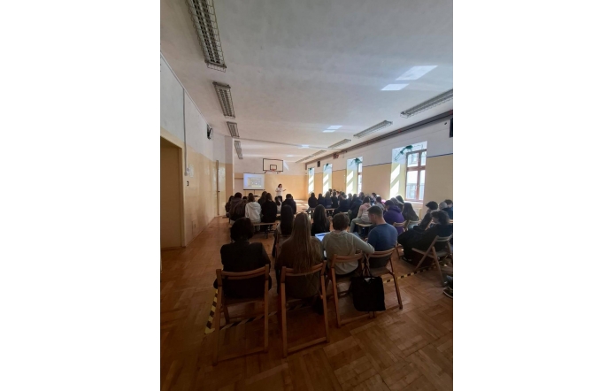Zdjęcie przedstawia salę szkolną z grupą uczniów siedzących na krzesłach ustawionych w rzędach. Uczniowie są zwróceni w stronę ekranu, na którym wyświetlana jest prezentacja. Po lewej stronie widoczne okna, na ścianach dekoracje.