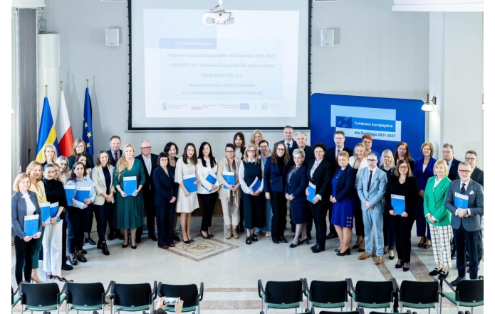 Grupa kilkudziesięciu osób pozuje do wspólnego zdjęcia w jasnej sali konferencyjnej, wiele z nich trzyma niebieskie teczki. W tle widoczne są flagi, ekran z prezentacją o Funduszach Europejskich oraz baner informacyjny.