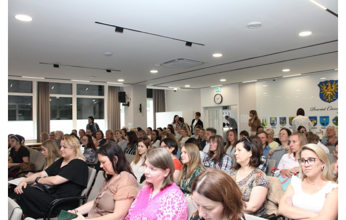 Duża grupa osób siedzi w rzędach w jasnej sali konferencyjnej. Większość uczestników jest zwrócona w jednym kierunku. W tle widoczne są herby gmin oraz herb Powiatu Cieszyńskiego.