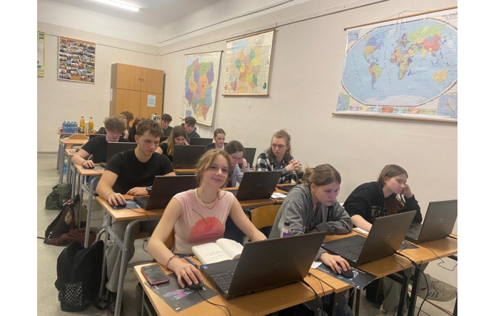 Uczniowie siedzą w sali lekcyjnej przy biurkach z laptopami, skupieni na pracy. Na ścianach wiszą mapy świata i Afryki. W tle stoją butelki z napojami. Jedna z uczennic uśmiecha się do obiektywu.