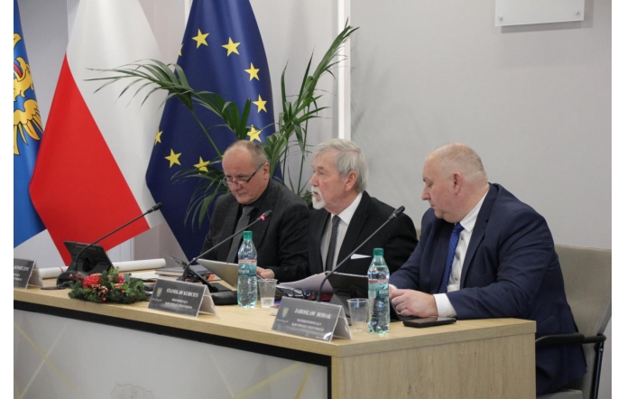Trzech mężczyzn siedzi za stołem prezydialnym z mikrofonami, dokumentami i butelkami wody. W tle widać flagi, roślinę oraz jasną salę obrad.