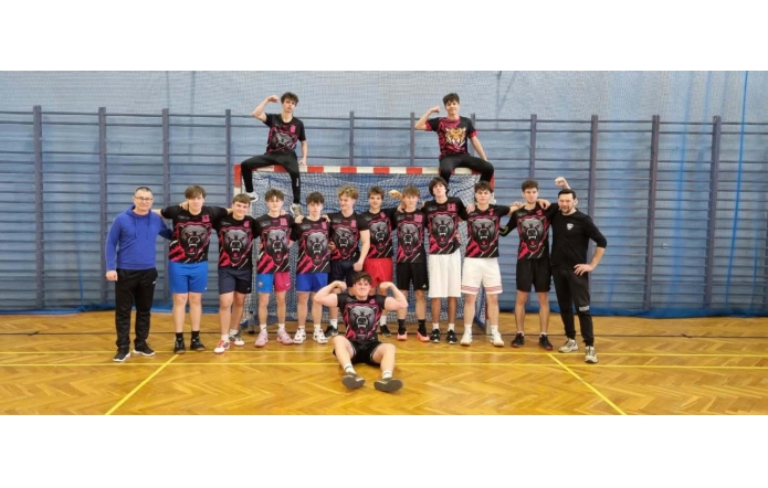 Hala sportowa z drewnianą podłogą i bramką w tle. Grupa nastolatków w czarno-czerwonych koszulkach z grafiką niedźwiedzia pozuje do zdjęcia razem z dwoma dorosłymi mężczyznami stojącymi po bokach. Dwóch chłopaków siedzi na górnej belce bramki, a jeden siedzi na podłodze z przodu i pokazuje zgięte ręce.