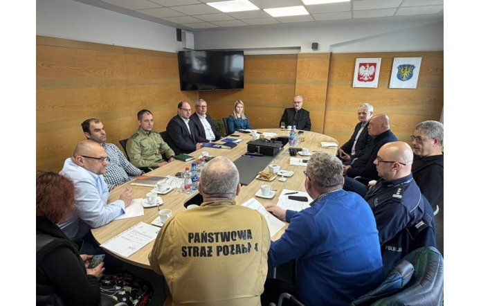 Grupa osób siedzących przy stole konferencyjnym podczas spotkania, z dokumentami, kawą i wodą, w tle ściana z herbami i ekranem.