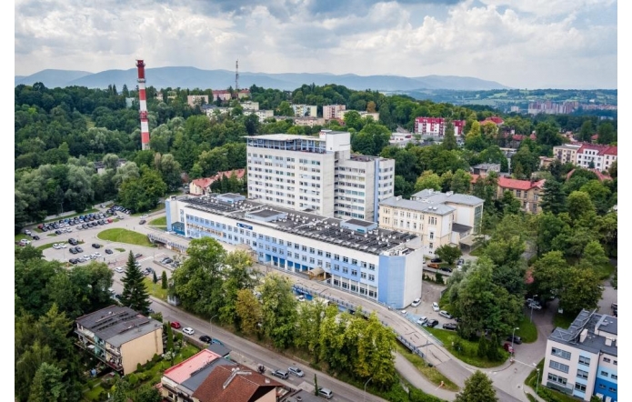 Rozległy kompleks budynków otoczony zielenią, widziany z lotu ptaka. Wokół znajdują się drogi, parkingi i zabudowa miejska, a w tle widoczne są wzgórza i góry.