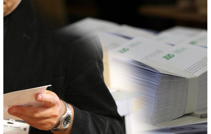Mężczyzna w ciemnym ubraniu trzyma w dłoni dokument. Obok znajduje się duży stos zadrukowanych przesyłek z logo ZUS, spiętych taśmą pakunkową, gotowych do wysyłki lub dystrybucji.