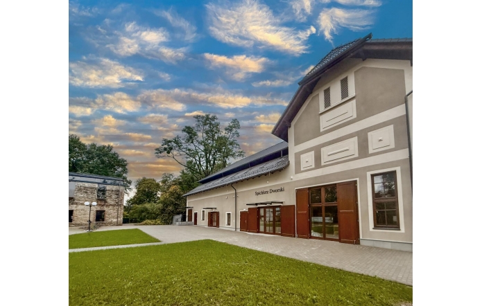 Budynek Spichlerza Dworskiego z drewnianymi okiennicami i jasną elewacją. Przed obiektem znajduje się zielony trawnik i utwardzony plac, a w tle widoczne są drzewa oraz efektowne, zachmurzone niebo.