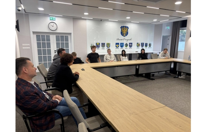 Grupa młodzieży i dorosłych siedzących przy dużym stole konferencyjnym, w jasnej sali.
