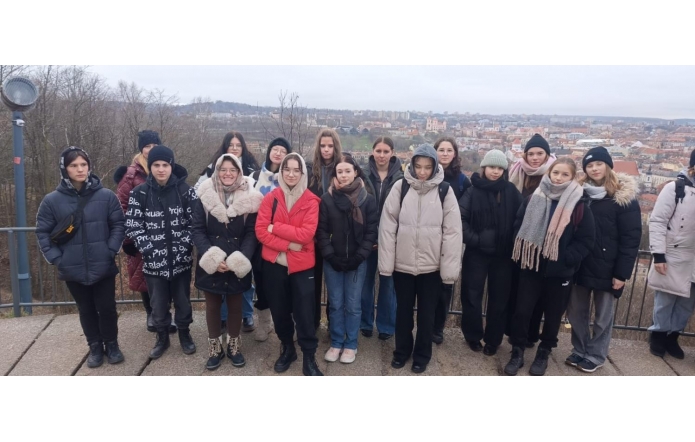Grupa młodych ludzi stoi na tle panoramicznego widoku Wilna, widocznego w oddali. Uczestnicy ubrani są w zimowe kurtki, szaliki i czapki, a ich twarze wyrażają skupienie i spokój. W tle widać czerwone dachy budynków, kościoły oraz rozciągający się krajobraz z drzewami i miejskimi uliczkami.
