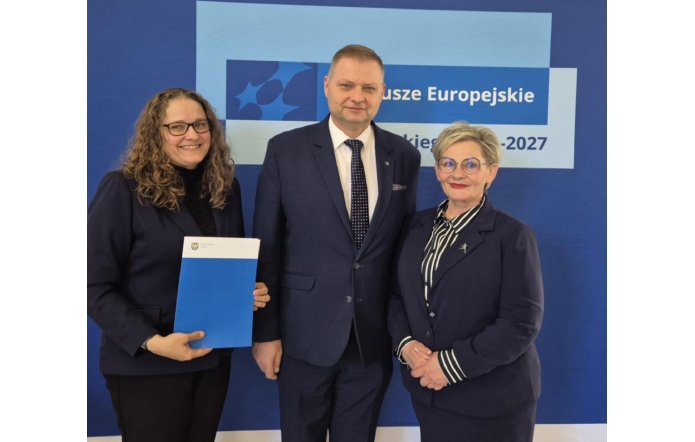 Trzy osoby w eleganckich strojach (od lewej Agnieszka Baszczyńska, dyrektor PCPR w Cieszynie, od prawej Beata Macura, Członkini Zarządu Powiatu Cieszyńskiego, w środku Rafał Adamczyk, Członek Zarządu Województwa Śląskiego) stoją na tle niebieskiej ścianki z napisem „Fundusze Europejskie 2021–2027”. Agnieszka Baszczyńska trzyma teczkę z logo województwa śląskiego. Wszyscy uśmiechnięci, pozują do wspólnego zdjęcia.