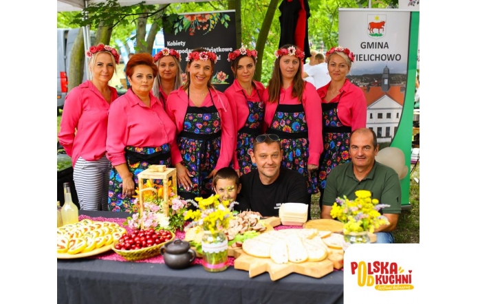 Grupa kobiet w różowych koszulach i kwiecistych fartuchach, z wiankami na głowach, stoi za stołem pełnym tradycyjnych potraw. Na stole widać chleb, wędliny, wiśnie i kwiaty. Obok nich siedzi dwóch mężczyzn i chłopiec. Scena odbywa się na tle zielonego parku.