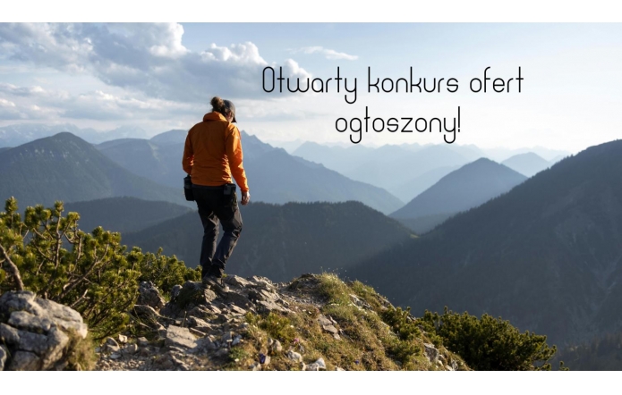 Grafika przedstawia osobę w pomarańczowej kurtce i czarnych spodniach stojącą na skalistym szczycie, patrzącą na widoczne w oddali pasma górskie. Na jasnym niebie ponad szczytami gór widnieje czarny napis „Otwarty konkurs ofert ogłoszony!”.