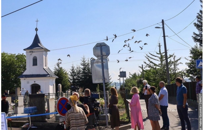 Grupa osób stoi przy ogrodzeniu w pobliżu niewielkiej kaplicy. Nad drogą przelatuje stado ptaków, a obok ustawiony jest sprzęt filmowy i znaki drogowe.
