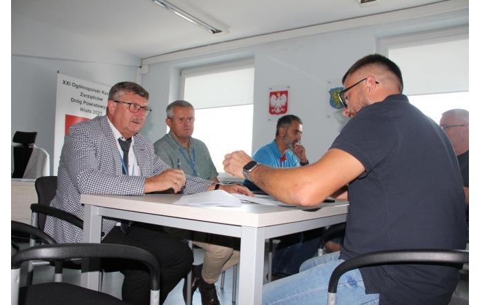 Na zdjęciu widać grupę mężczyzn siedzących przy stole w biurze. Dwóch z nich siedzi po prawej stronie stołu, a trzech po lewej Jerzy Pilch, Członek Zarządu, Jarosław Kalinik, Dyrektor Powiatowego Zarządu Dróg Publicznych w Cieszynie, Ryszard Krzywoń, Zastępca Dyrektora Powiatowego Zarządu Dróg Publicznych w Cieszynie. Pierwszy mężczyzna po lewej stronie: Jerzy Pilch ma na sobie marynarkę w kratkę i okulary, trzyma długopis i patrzy na dokumenty leżące na stole. Drugi mężczyzna: Jarosław Kalinik siedzący obok niego ma na sobie szarą koszulę z długim rękawem. Trzeci mężczyzna: Ryszard Krzywoń ma na sobie niebieską koszulę. Po prawej stronie stołu siedzi mężczyzna w ciemnej koszulce z krótkim rękawem, również z okularami. Za nim widać kolejnego mężczyznę w okularach. Na ścianie wiszą dwa godła oraz tablica z napisem.