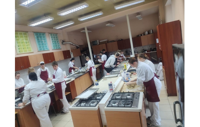Grupa uczniów w białych strojach kucharskich i bordowych fartuchach pracuje w szkolnej pracowni gastronomicznej. Część osób wyrabia ciasto przy blatach, inni przygotowują składniki i korzystają z piekarników oraz kuchenek gazowych. Pomieszczenie wyposażone jest w drewniane szafki, metalowe blaty i oświetlenie sufitowe.