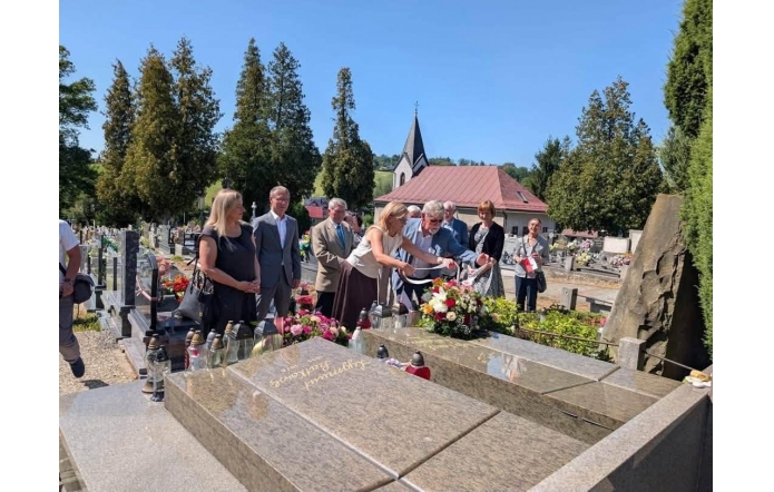 Grupa osób stoi przy grobie na cmentarzu. Dwie osoby na pierwszym planie układają kwiaty i znicze, a pozostali stoją obok. W tle widoczne są nagrobki, drzewa i budynek z wieżyczką.