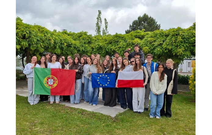 Grupa młodzieży pozuje do wspólnego zdjęcia z flagami Portugalii, Polski i Unii Europejskiej. Zielone otoczenie oraz pochmurne niebo.