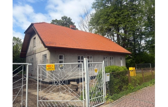 Remontowany budynek. Przed wejściem znajduje się furtka, tablica informacyjna oraz zadbany ogród z krzewami i kwiatami.