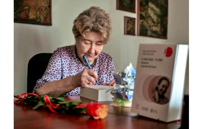 prof. Krystyną Heską-Kwaśniewicz, ubrana w elegancką, wzorzystą bluzkę, siedzi przy biurku i podpisuje książkę. Na biurku widoczna jest pomarańczowa, przystrojona wstążką jedna róża, obok niej na stojaku stoi książka. W tle, na ścianie widać wiszące obrazy.