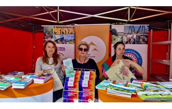 Trzy kobiety – od lewej: Aneta Brożkowska, główna specjalistka w Biurze ds. Kultury, Sportu, Turystyki i Promocji Powiatu, Beata Macura, Członkini Zarządu, Barbara Jurkiewicz, Kierowniczka Biura ds. Kultury, Sportu, Turystyki i Promocji Powiatu, stoją za stoiskiem pełnym kolorowych ulotek i broszur. Uśmiechają się, trzymając torby i materiały promocyjne. Tło ozdobione jest plakatami z motywami turystycznymi. Jasne, soczyste kolory tworzą przyjazną atmosferę.