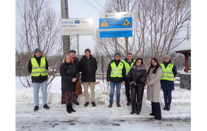Grupa dziewięciu osób stoi na ośnieżonym poboczu drogi, pod tablicami informującymi o realizowanej inwestycji drogowej w ramach programu Interreg Polska–Słowacja oraz o prowadzonych pracach budowlanych. Wśród zebranych znajdują się przedstawiciele Powiatu i PZDP, wicewójt Istebnej oraz szef firmy Drogród. Część osób ma na sobie kamizelki odblaskowe, pozostali ubrani są w zimowe płaszcze i czapki. W tle widoczne są drzewa bez liści i zimowy krajobraz.