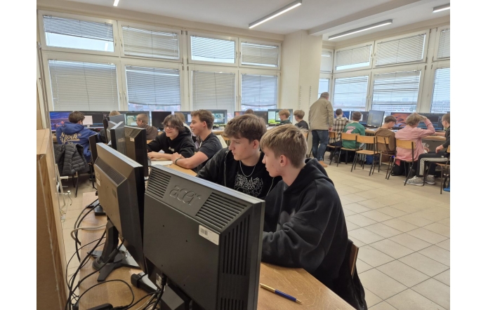 Uczniowie siedzą przy stanowiskach komputerowych w pracowni i pracują przy monitorach. Część osób siedzi parami, a w tle widoczne są kolejne stanowiska oraz osoba stojąca między rzędami komputerów.