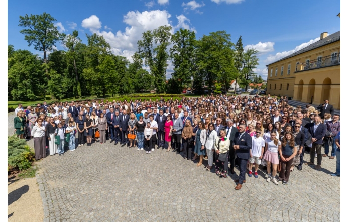 Uczestnicy wydarzenia pozują do wspólnego zdjęcia na dziedzińcu przed budynkiem. Na fotografii widoczna jest liczna grupa osób zgromadzonych na otwartej przestrzeni. W tle znajdują się drzewa, park oraz zabudowania.