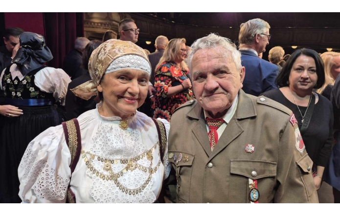 Kobieta w stroju regionalnym stoi obok mężczyzny w mundurze z odznaczeniami. Oboje patrzą w stronę aparatu. W tle widoczne są liczne osoby zgromadzone we wnętrzu sali.