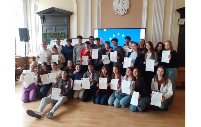 Grupa młodzieży w jasnej sali z drewnianą podłogą pozuje z certyfikatami. W tle ekran z flagą UE i godło Polski. Wszyscy uśmiechnięci, ubrani na luzie, stoją i siedzą w kilku rzędach, świętując wspólne osiągnięcie.