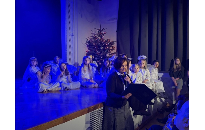 Janina Żagan Starosta Cieszyński przemawia do mikrofonu przy scenie, trzymając teczkę. Na scenie siedzą dzieci w białych strojach, a w tle widać choinkę i niebieskie oświetlenie.