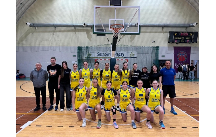 Drużyna koszykarska dziewcząt ustawiona do wspólnego zdjęcia na hali sportowej, w żółtych strojach, z trenerami i sztabem w tle oraz koszem do gry i tablicą wyników nad nimi.