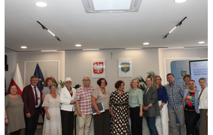 Grupa uczestników spotkania pozuje do wspólnego zdjęcia w sali sesyjnej. Osoby stoją w jednym rzędzie, część z nich trzyma dokumenty. 