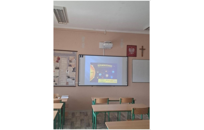 Zdjęcie przedstawia salę lekcyjną. Na ścianie wyświetlony jest slajd z prezentacji „Dermokosmos”. Przed ścianą ustawione są ławki i krzesła. Po lewej stronie widoczne plakaty edukacyjne, po prawej tablica.