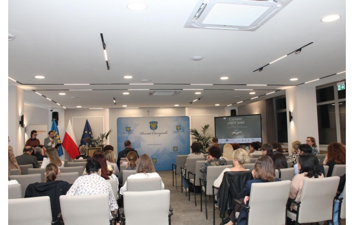 Sala sesyjnej z publicznością siedzącą na krzesłach. Z przodu stoją prowadzący przy mównicy, obok flagi, a w tle widać ekran z prezentacją i planszę Powiatu Cieszyńskiego.