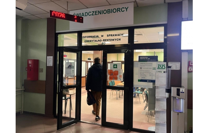 Wejście do pomieszczenia w ZUS, związanego z obsługą świadczeniobiorców. Nad drzwiami widnieje elektroniczny wyświetlacz z numerkami, wskazujący kolejność obsługi. Mężczyzna w ciemnej kurtce i jasnych spodniach przechodzi przez przeszklone drzwi, trzymając w ręku torbę. W środku ustawione są biurka, krzesła i tablice informacyjne. Na ścianach widoczne są plakaty i instrukcje dla klientów.