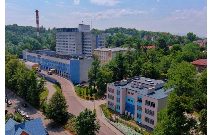 Widok z góry na kompleks Szpitala Śląskiego w Cieszynie. Wśród zieleni znajdują się budynki szpitalne, droga dojazdowa oraz zaparkowane samochody. Tło tworzą drzewa, zabudowania miejskie i jasne niebo.