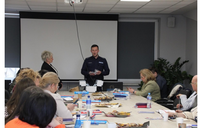 Policjant prowadzi spotkanie w sali konferencyjnej, mówiąc do grupy osób siedzących przy stole. Przed nim ekran projekcyjny, na stole materiały, przekąski i napoje. Uczestnicy słuchają, notują lub przeglądają broszury.